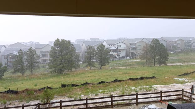 A vivid image capturing a heavy hailstorm over a suburban neighborhood. The storm pelts down on houses, trees, and an open field, creating a dramatic scene of intense weather conditions.