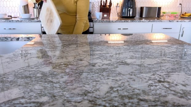 A woman in a yellow sweater slices a fresh watermelon on a cutting board in a modern kitchen. The kitchen features white cabinetry, a hexagonal tile backsplash, and various cooking utensils on the countertop.