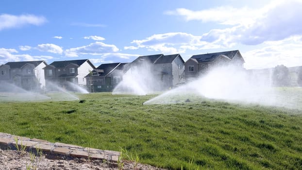 A suburban neighborhood scene featuring automatic sprinklers watering a lush green lawn in front of modern homes. The sprinklers create a misty effect, emphasizing the freshness and care of the landscape.