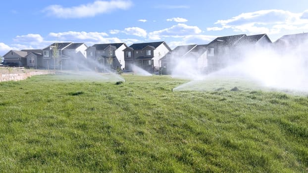 A suburban neighborhood scene featuring automatic sprinklers watering a lush green lawn in front of modern homes. The sprinklers create a misty effect, emphasizing the freshness and care of the landscape.