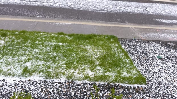 A lawn and rock garden blanketed in hail following a storm, with the green grass and small shrubs peeking through the layer of ice. The contrast between the white hail and the greenery creates a striking visual effect.