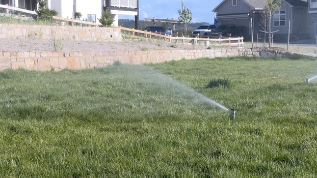 A suburban neighborhood scene featuring automatic sprinklers watering a lush green lawn in front of modern homes. The sprinklers create a misty effect, emphasizing the freshness and care of the landscape.