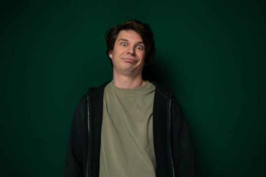 A man is standing upright in front of a bright green wall, looking towards the camera. He appears relaxed and casual in attire, with hands in pockets.
