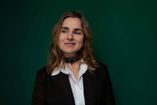 A woman is standing in front of a solid green wall, with a neutral expression on her face. She appears to be looking straight ahead, with her hands relaxed by her sides.