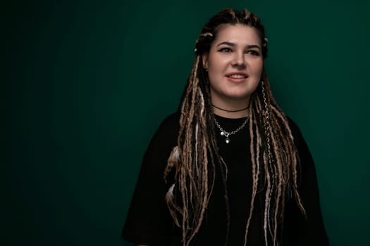 A woman with dreadlocks is standing in front of a bright green background. She is looking directly at the camera with a neutral expression. The womans hair is styled in long, thin, twisted dreadlocks, and she is wearing casual clothing.