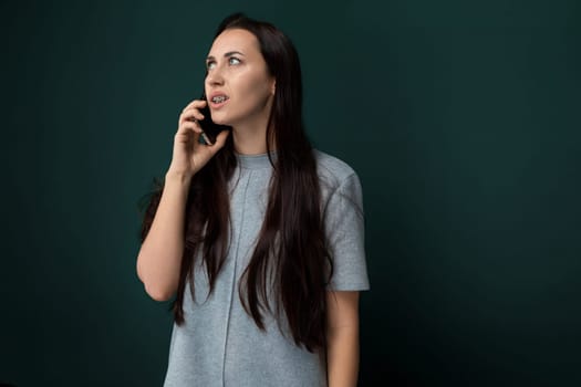 A woman is standing in front of a green wall while speaking on her cell phone. She appears engaged in the conversation as she gestures slightly with her free hand.