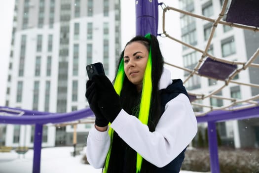 A woman stands in front of a basketball hoop, holding a cell phone up. She appears to be taking a photo or video of the hoop. The setting is a basketball court.