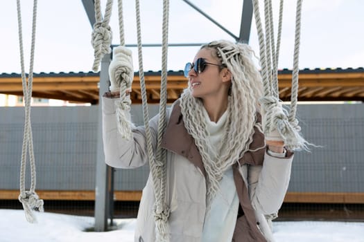 A woman with white dreadlocks is sitting on a swing, moving back and forth with her feet off the ground. The swing is in motion, and the womans hair is flowing behind her.