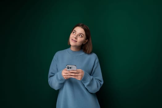A woman wearing a blue sweater is seen holding a cell phone in her hand, appearing engaged with the device. She stands in a casual pose, looking at the screen while surrounded by a simple background.