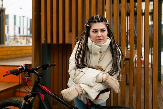 A woman with dreadlocks standing next to a stationary bicycle on a city street. She is wearing casual clothing and appears to be taking a break from cycling.