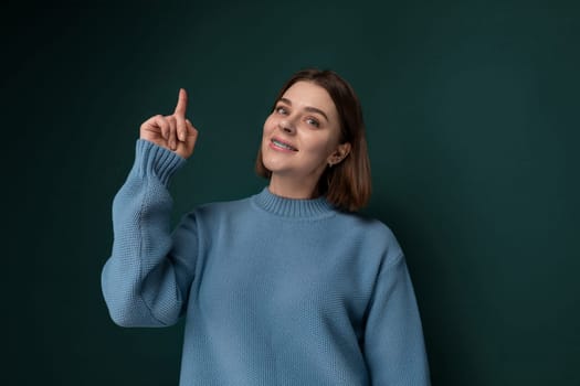 A woman wearing a blue sweater is pointing at an unspecified object or direction. She appears to be gesturing with intent and focus, possibly indicating something of importance or interest. The background is undefined, keeping the viewers attention on the woman and her action.