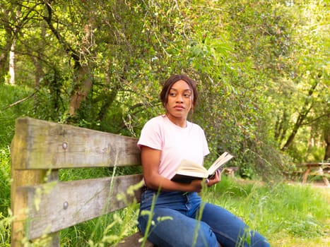 Young black woman sitting on a park bench reading a book.