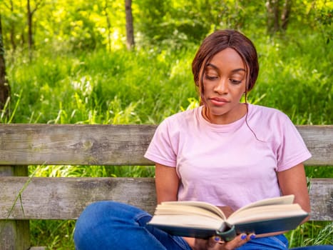 Young black woman sitting on a park bench reading a book.