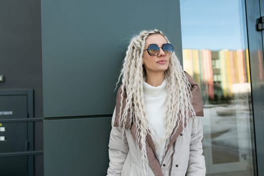 A woman with white dreadlocks is seen standing in front of a building, looking directly at the camera with a confident expression. The building behind her is tall and modern, with large windows and a sleek design.