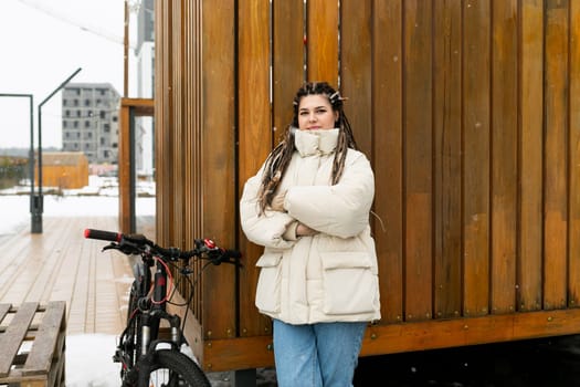 A woman is standing next to a bike in front of a building. She is looking towards the camera, with the bike beside her. The building behind her is tall and modern, adding to the urban setting of the scene.