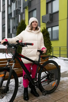 A woman is standing next to a bicycle in front of a wooden bench. She appears to be taking a break or resting momentarily. The scene is simple yet urban, with a focus on the interaction between the woman, the bike, and the bench.