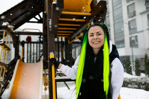 A woman with vibrant green hair stands confidently in front of a colorful playground, looking towards the camera. She is surrounded by swings, slides, and other playground equipment.