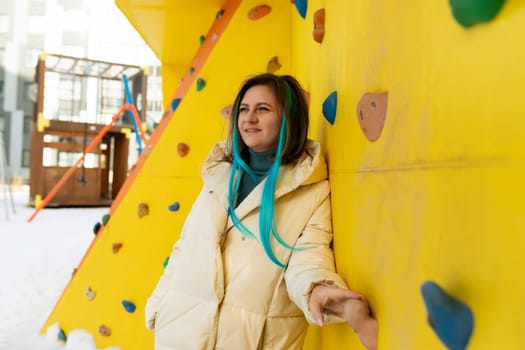 A woman in athletic attire stands confidently next to a bright yellow climbing wall, ready to tackle the challenging ascent. She appears focused and determined, showcasing strength and agility in this sporty setting.