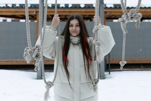 A woman in warm clothing stands on a swing covered in snow, holding onto the chains for balance. The snow-covered ground and trees in the background indicate a winter setting.