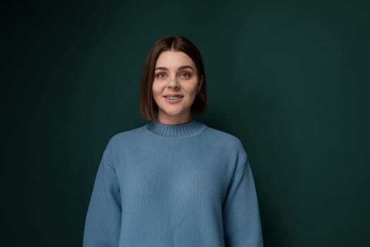 A woman standing in front of a plain green wall, looking directly at the camera. She appears relaxed and confident, with a neutral expression. The background is unadorned and focuses the viewers attention on the woman.