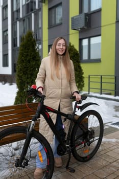 A woman wearing _______ is standing next to a bicycle in front of a wooden bench. She appears to be in a park or outdoor setting, with trees in the background.