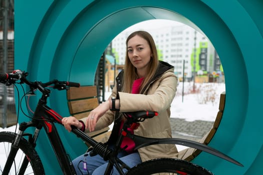 A woman wearing a red jacket sitting on a wooden bench next to a blue bicycle. The setting is an urban park, with trees and buildings in the background.