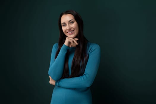 A woman in a stylish blue dress confidently poses for a photograph, standing with one hand on her hip and flashing a bright smile. She appears elegant and poised.