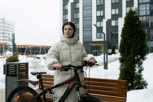 A woman stands next to a bike in a snow-covered landscape. She is dressed warmly, looking around the snowy scenery. The bike is parked on the snowy ground, with snowflakes falling gently around them.
