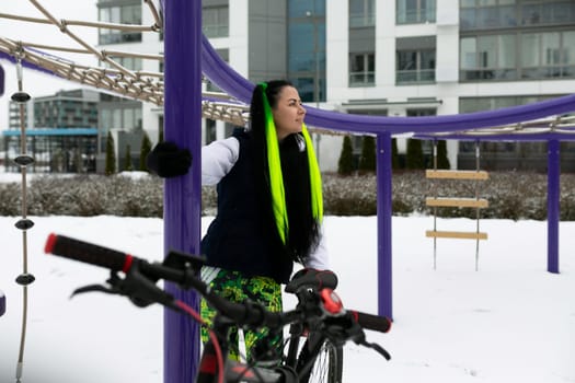 A woman in winter clothing stands next to a bicycle parked in the snow-covered ground. The scene depicts a cold winter day with the woman taking a break from cycling.