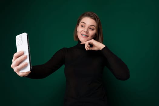 A woman is standing outdoors, holding her cell phone at arms length, and smiling as she takes a selfie.