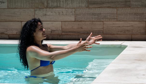 Smiling latin pretty woman with open arms in a pool in summer holidays.