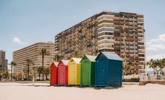 Multicolored beach huts with tall residential buildings and palm trees in the background, creating a vibrant coastal scene. Sant Antoni beach, Cullera, Spain