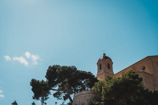 The tower of a historic church surrounded by trees against a clear blue sky. Cullera Castle, Spain