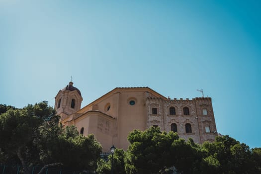 A historic church building with trees in the foreground and a clear blue sky above. Cullera Castle, Spain