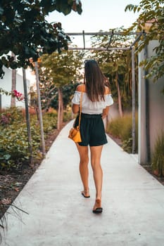 Woman walking on a paved path surrounded by lush greenery. The relaxed setting features trees and plants, capturing a serene moment in a beautiful, natural environment. The woman s back is visible.