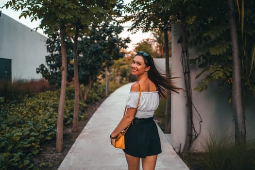 smiling woman strolling on a garden path surrounded by trees and greenery at sunset. The warm light creates a tranquil atmosphere, capturing a relaxed, joyful moment in a natural setting