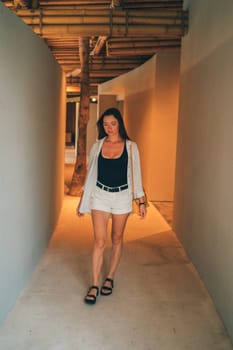 A woman walking through a modern architectural passageway. The minimalistic design features smooth walls and bamboo ceilings, creating a stylish, serene atmosphere. The woman s expression is calm and composed.