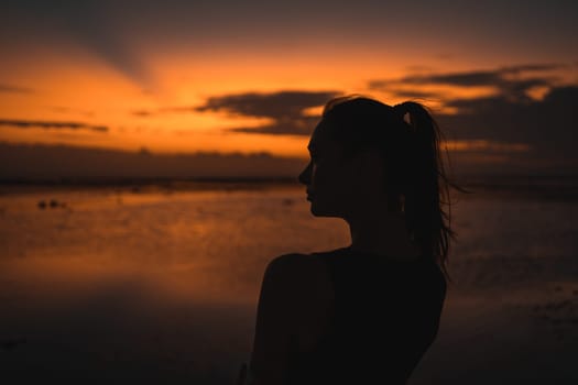 Silhouette of a woman at sunset on the beach, facing the ocean. The vibrant sky and calm water create a serene, contemplative atmosphere, capturing a peaceful moment in nature.