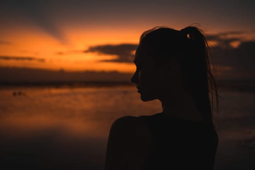 Silhouette of a beautiful woman standing at sunset by the ocean. The vibrant sky and calm water create a serene, contemplative atmosphere, capturing a peaceful moment in nature.