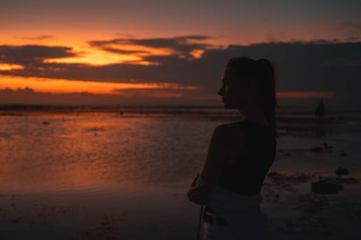 Woman contemplating the sunset by the sea, captured in silhouette. The vibrant sky and reflective water create a serene, contemplative scene, emphasizing the beauty and tranquility of the moment.