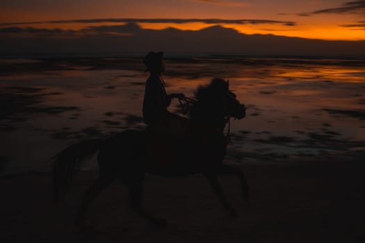 Horse unrecognized rider on the beach during sunset, captured in silhouette against a vibrant sky. The scene reflects on the wet sand, creating a picturesque, serene moment by the ocean.