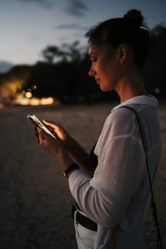 Profile of a woman using a smartphone at dusk on the beach. The soft lighting captures her focused expression, blending technology and natural evening ambiance. Film noise imitation added.