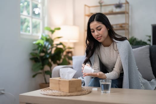 Young woman with the flu, wrapped in a blanket, taking medication while sitting on a sofa, demonstrating the concept of illness and flu treatment