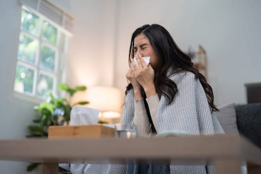 Young woman with a cold and runny nose sneezing into a tissue, wrapped in a blanket while sitting on a sofa, demonstrating the concept of cold and flu relief