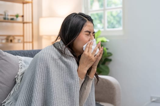 Young woman with cold symptoms sneezing into a tissue, wrapped in a blanket while sitting on a sofa, demonstrating the concept of cold and flu relief