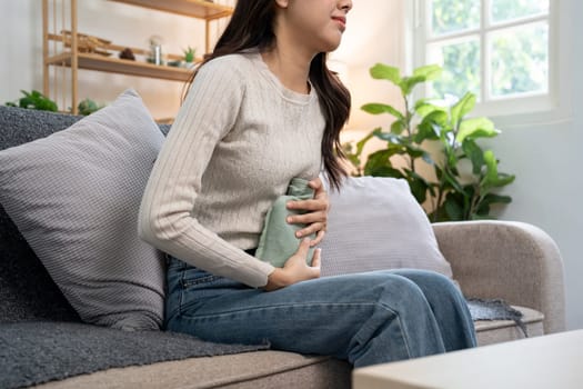 Young woman using a heat pad to relieve menstrual cramps while sitting on a sofa in a cozy living room, demonstrating the concept of menstrual pain management