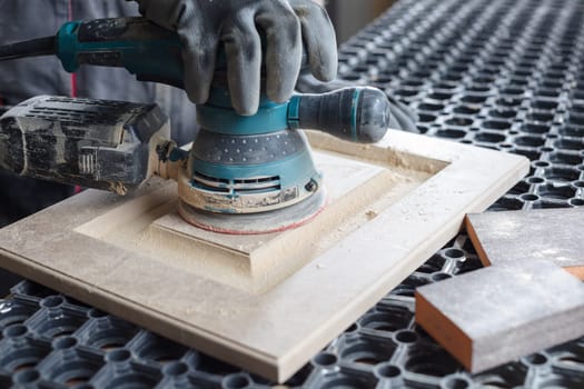 A male carpenter sands wooden furniture facades with a sanding machine at a furniture production factory, wood processing concept