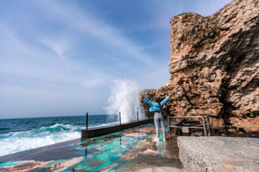 A woman in a blue jacket stands on a rock above a cliff above the sea and looks at the raging ocean. Girl traveler rests, thinks, dreams, enjoys nature. Peace and calm landscape, windy weather