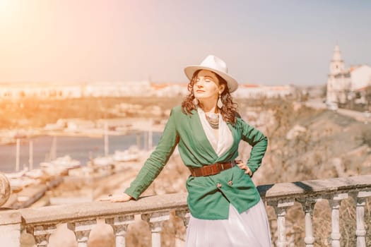 Woman walks around the city, lifestyle. Happy woman in a green jacket, white skirt and hat is sitting on a white fence with balusters overlooking the sea bay and the city