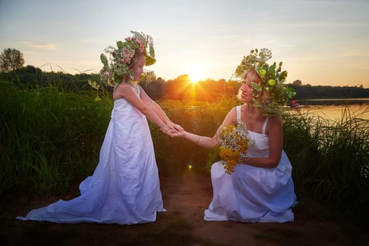 Mother and daughter in white sundresses, wreaths of flowers and bouquet in nature evening at sunset . Family celebrate Slavic Holiday of Ivan Kupala